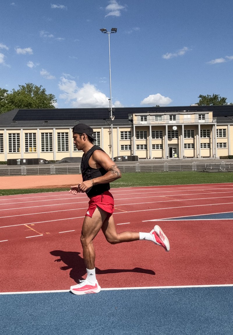 Αθλητής που φοράει κόκκινα σορτς και μαύρο τοπ τρέχει σε εξωτερική πίστα με ένα κτήριο και γαλάζιο ουρανό στο φόντο.