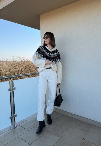 Mujer con suéter estampado, pantalones blancos, botas negras y gafas de sol de pie en un balcón sosteniendo un bolso negro.