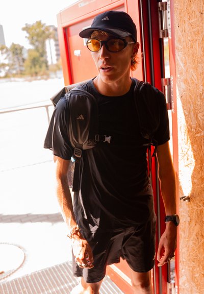 Camiseta y pantalones cortos deportivos negros, usando una gorra negra y gafas de sol con tintes amarillos. Mochila con correas, de pie frente a una puerta. Iluminación brillante.