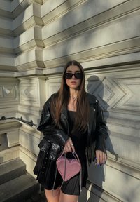 Svart faux läder oversized jacka, svart klänning med vertikala ränder, rosa hjärtformad handväska med kedjesnodd, solglasögon.