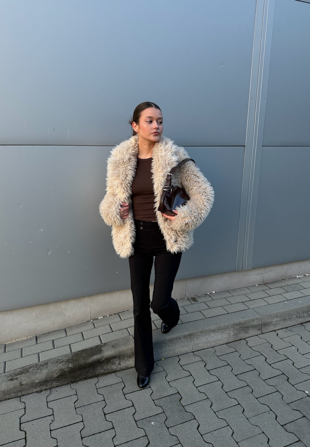 Jakna iz umetnega krzna krem barve, rjava oprijeta majica, črne razširjene hlače in črne pete, z rjavo torbico, stoji ob sivi steni.