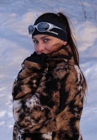 Bruin en zwart patroon fleecejack met lange mouwen, gedragen met zwarte handschoenen en hoofdband. Sneeuwerige achtergrond met zachte, vage textuur.