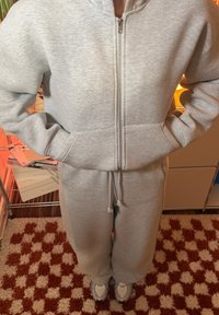 Sweat à capuche gris zippé et pantalon assorti en matériau doux, avec poches latérales et cordons à la taille. Les chaussures sont blanches avec des accents gris.