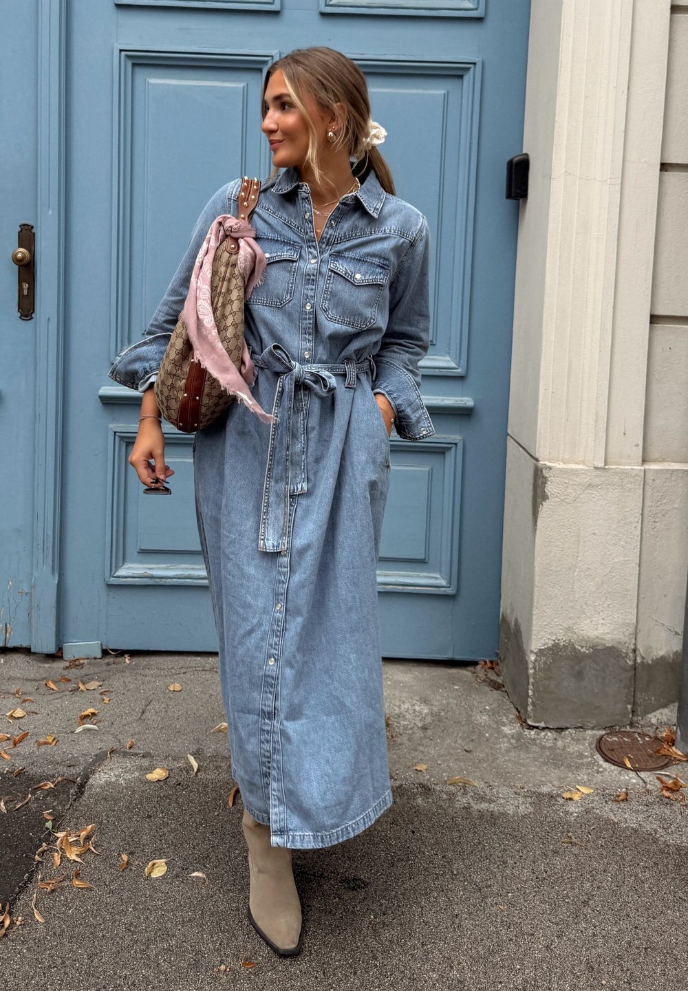 Vestito camicia in denim, azzurro chiaro, maniche lunghe, vita con cintura, apertura frontale con bottoni. Accessoriato con una borsa a fantasia e una sciarpa leggera.