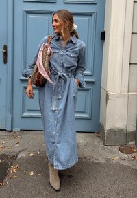 Vestito camicia in denim, azzurro chiaro, maniche lunghe, vita con cintura, apertura frontale con bottoni. Accessoriato con una borsa a fantasia e una sciarpa leggera.