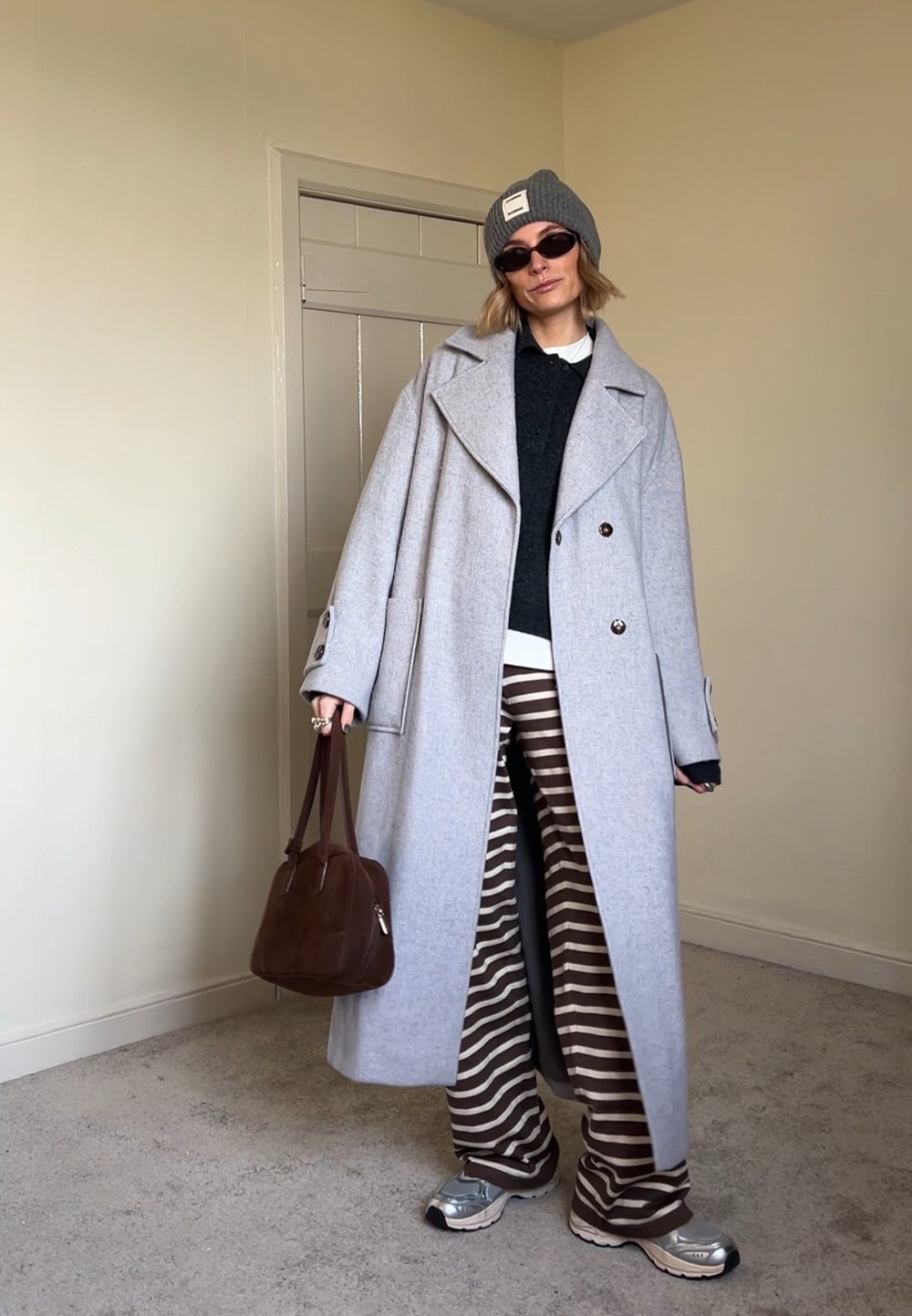 Light grey wool coat with large buttons, green knit jumper, striped brown and white trousers, grey beanie, small brown bag, and silver trainers.