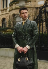 Long manteau trench-coat en cuir vert avec une taille ceinturée, un col superposé et des poches latérales. Sac à main noir avec des accents de quincaillerie dorée.