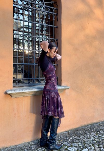 Vestido floral en púrpura oscuro con un corsé ajustado y dobladillo acampanado. Combinado con botas de cuero negro hasta la rodilla. El fondo presenta una ventana de hierro forjado.