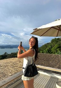 Musta nahkainen käsilaukku hopeaketjuhihnalla, valkoinen hihaton toppi ja vaalea röyhelöhelmainen hame. Seisoo puisella terassilla, jolta avautuu rannikkonäkymä.