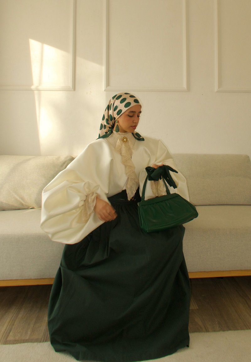 Femme assise sur un canapé beige, portant un haut blanc à manches bouffantes, une jupe vert foncé, un foulard à pois, tenant un sac à main vert assorti et des gants.