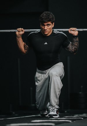 Hombre con tatuajes realizando una zancada con peso, sosteniendo una barra con pesas sobre los hombros, vestido con camisa negra y pantalones deportivos grises en un gimnasio.