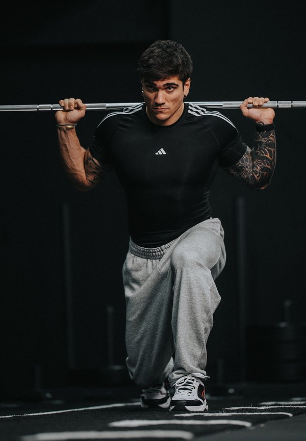 Tetovált férfi súlyozott kitörést végez, miközben egy súlyzórudat tart a vállán, fekete pólót és szürke melegítőnadrágot viselve az edzőteremben.