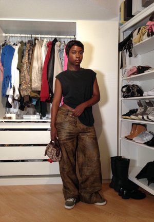 Top negro sin mangas, pantalones amplios marrón con estampado, sosteniendo un pequeño bolso marrón. Armario con varios artículos de ropa y zapatos al fondo.