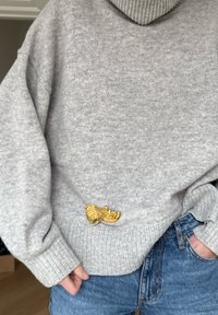 Personne portant un pull gris en laine avec une broche en forme de plume dorée épinglée près de la taille, assorti à un jean bleu et les mains dans les poches.