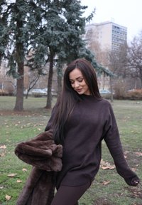 Žena v tmavom svetri a nohaviciach drží hnedý kožuch a kráča po tráve s stromami a budovami v pozadí počas neskorej jesene.