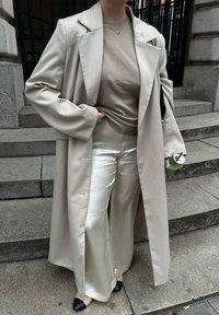 Personne portant un trench-coat beige, un pull taupe, un pantalon en satin crème, des ballerines beiges à bout noir, tenant une canette de boisson verte sur des marches en pierre.