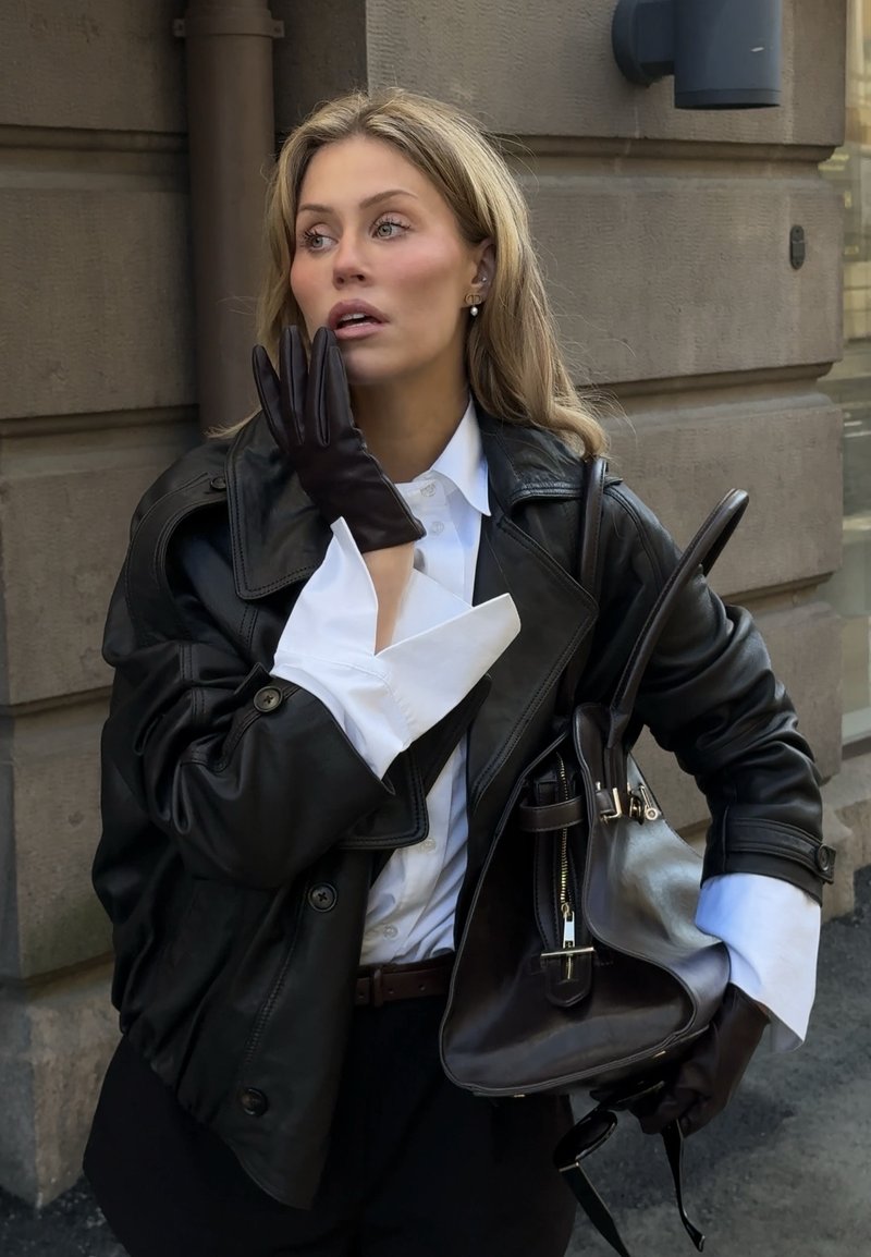 Kobieta w czarnej skórzanej kurtce i rękawiczkach trzyma pasującą torebkę, patrzy w bok z ręką blisko ust na tle kamiennego budynku.