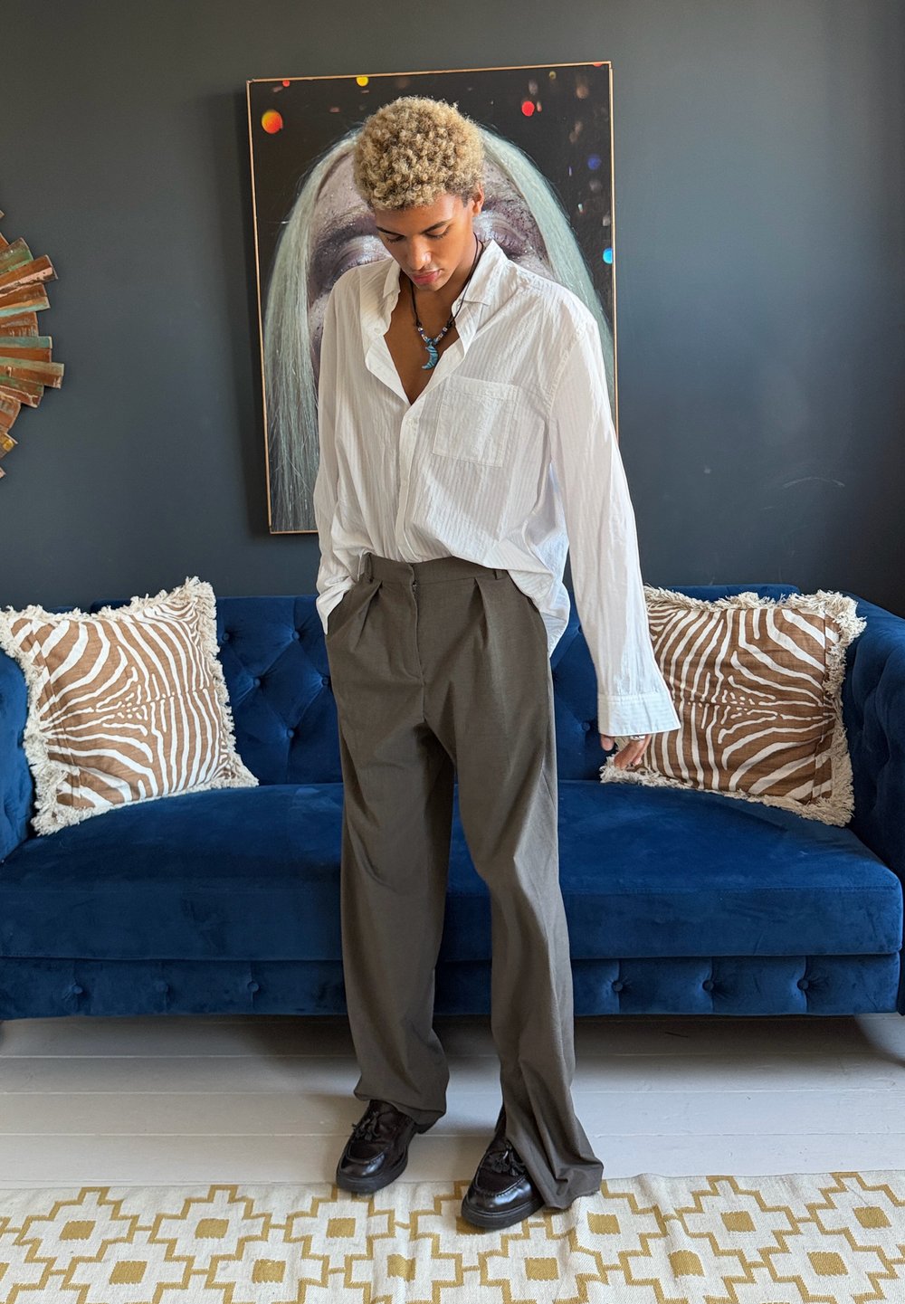 Jeune personne aux cheveux blonds bouclés portant une chemise blanche, un pantalon marron et des chaussures noires, se tenant devant un canapé bleu avec des coussins à motifs.