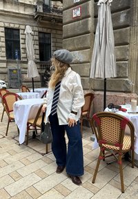 Hvit oversized skjorte, blå stripete slips, utsvingte denimbukser, grå beret og brune sko, plassert i en kafé med kurvstoler og bord.