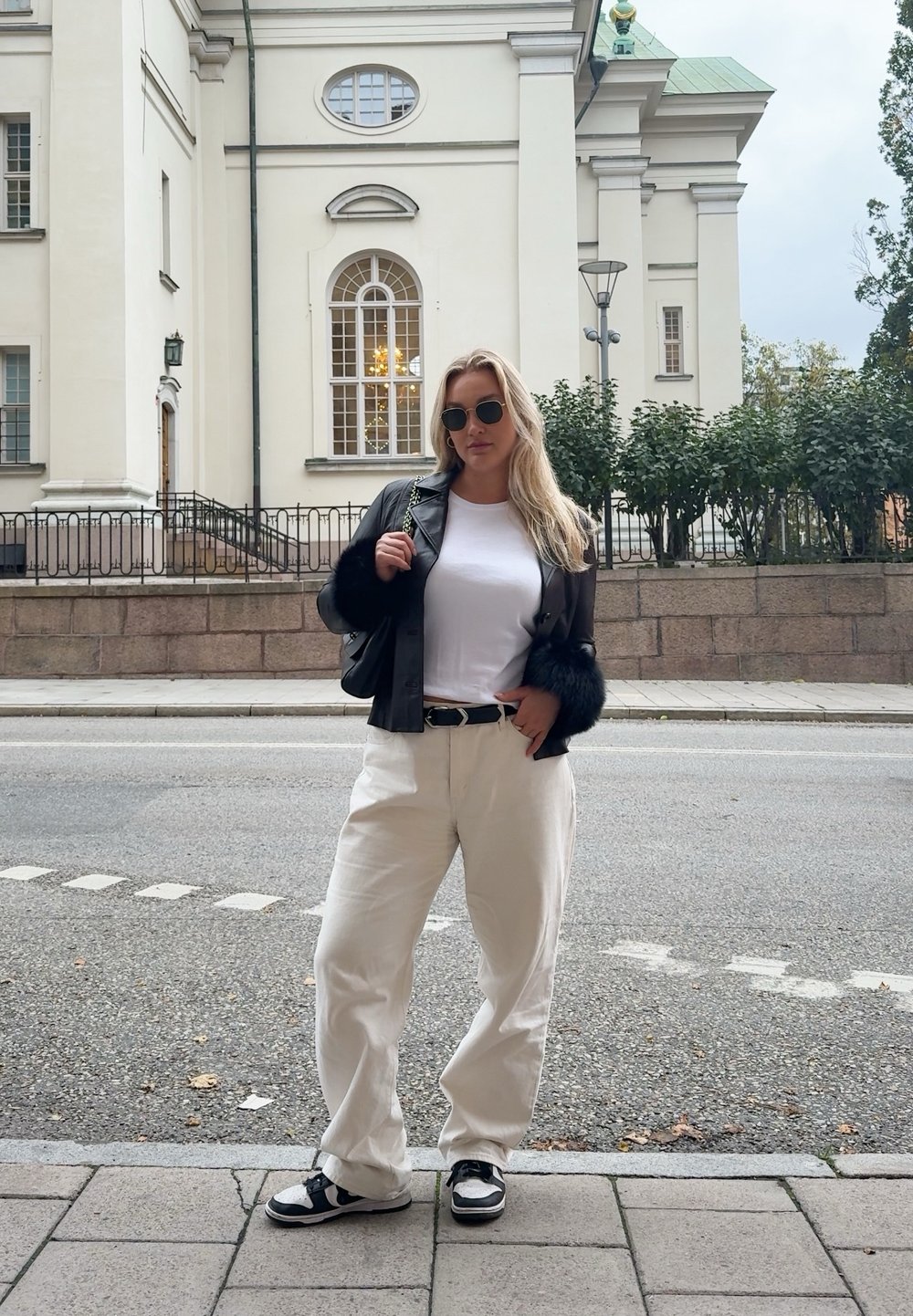 Black leather jacket with fur accents, white t-shirt, cream wide-leg trousers, and black trainers. The background features a light-coloured building and foliage.