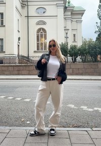 Black leather jacket with fur accents, white t-shirt, cream wide-leg trousers, and black trainers. The background features a light-coloured building and foliage.
