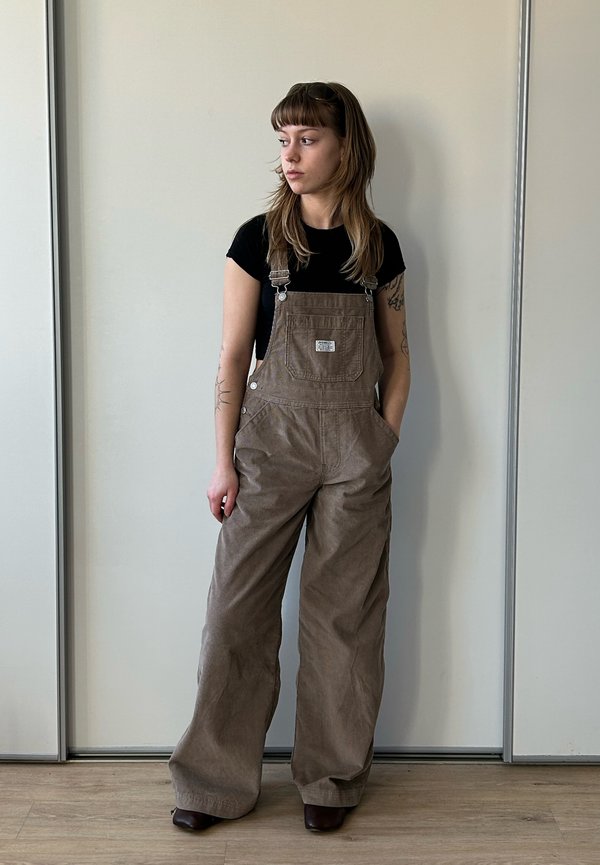 Jonge vrouw met lang haar kijkt naar links, draagt een bruine overall en een zwart T-shirt, staat tegen een effen lichte muur en houten vloer.