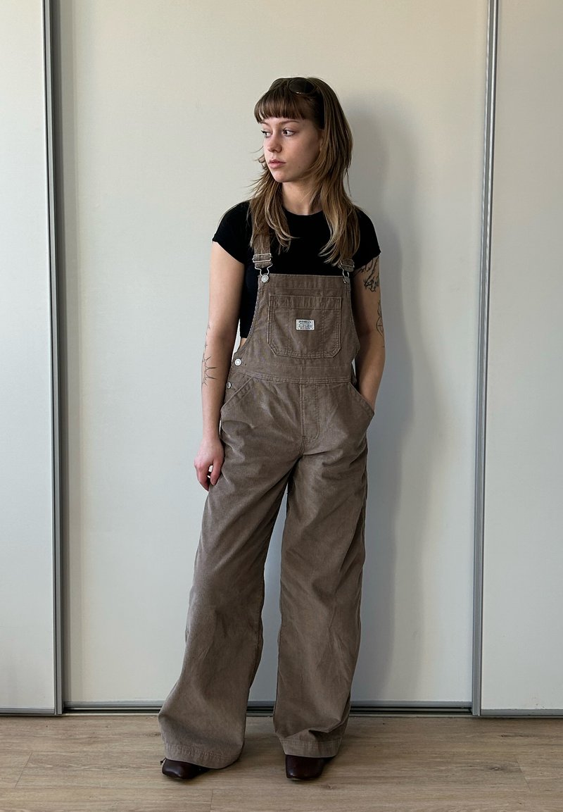 Jeune femme aux longs cheveux regarde à gauche, portant une salopette marron et un t-shirt noir, debout devant un mur clair uni et un sol en bois.