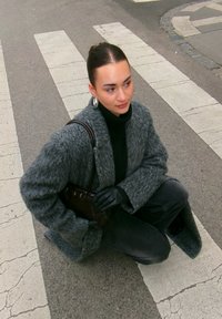 Manteau gris surdimensionné et duveteux, col roulé noir, gants, assis sur un passage pour piétons, tenant un petit sac à main noir, fond neutre avec des rayures blanches.