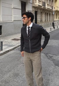 Veste zippée sombre avec col et poche avant, associée à un pantalon en velours côtelé beige ; un homme se tient dans une rue de la ville, les mains dans les poches.