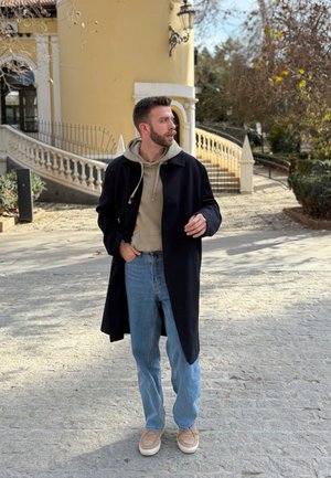 Abrigo negro sobre sudadera beige, jeans azul claro y calzado beige. De pie en un camino de piedras con un fondo de vegetación y arquitectura.