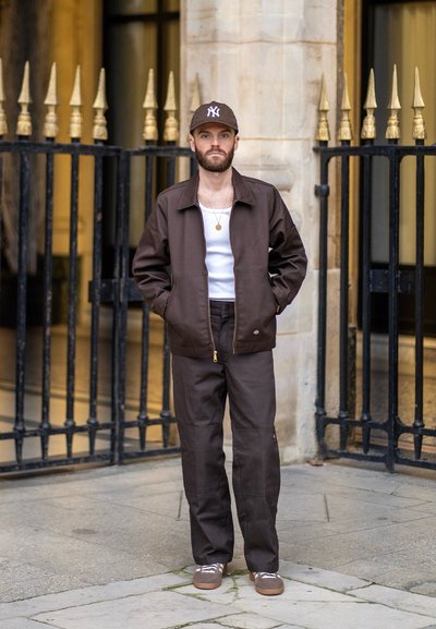 Chaqueta marrón con cremallera frontal, combinada con una camiseta sin mangas blanca y pantalones marrones. Gorro marrón con logo, zapatos marrones. Tejido texturizado.