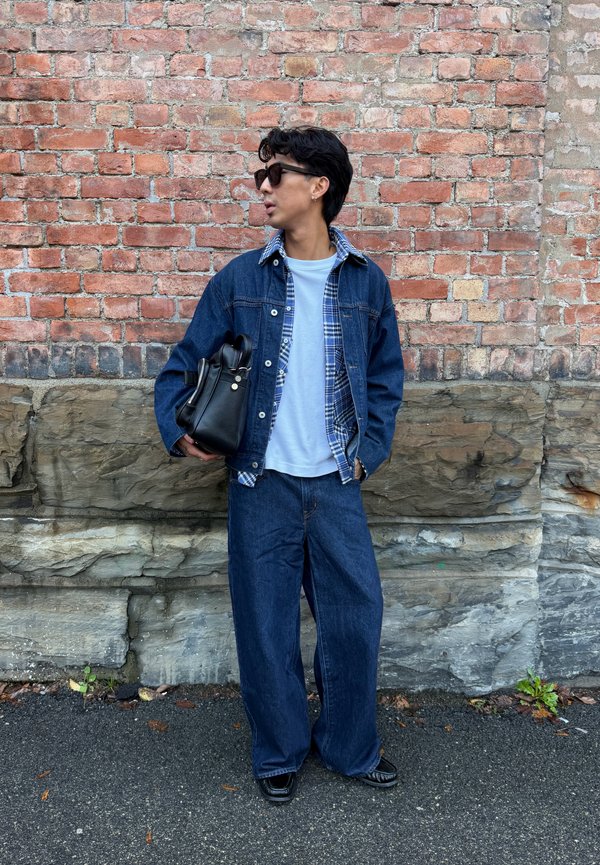 Una persona con gafas de sol, chaqueta vaquera y pantalones vaqueros está junto a una pared de ladrillo, sostiene un bolso negro y mira hacia un lado.