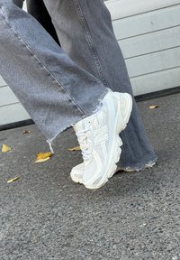 Ténis brancos em malha e sintético com uma sola robusta, apresentando um detalhe refletor e uma marca visível, combinados com jeans cinzentos desfiados.