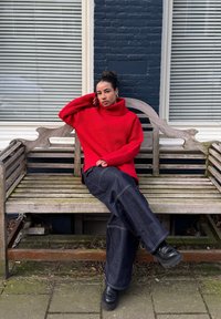 Maglione rosso a coste con collo alto, pantaloni di denim blu larghi e scarpe platee nere, seduto su una panchina di legno contro un muro di mattoni.