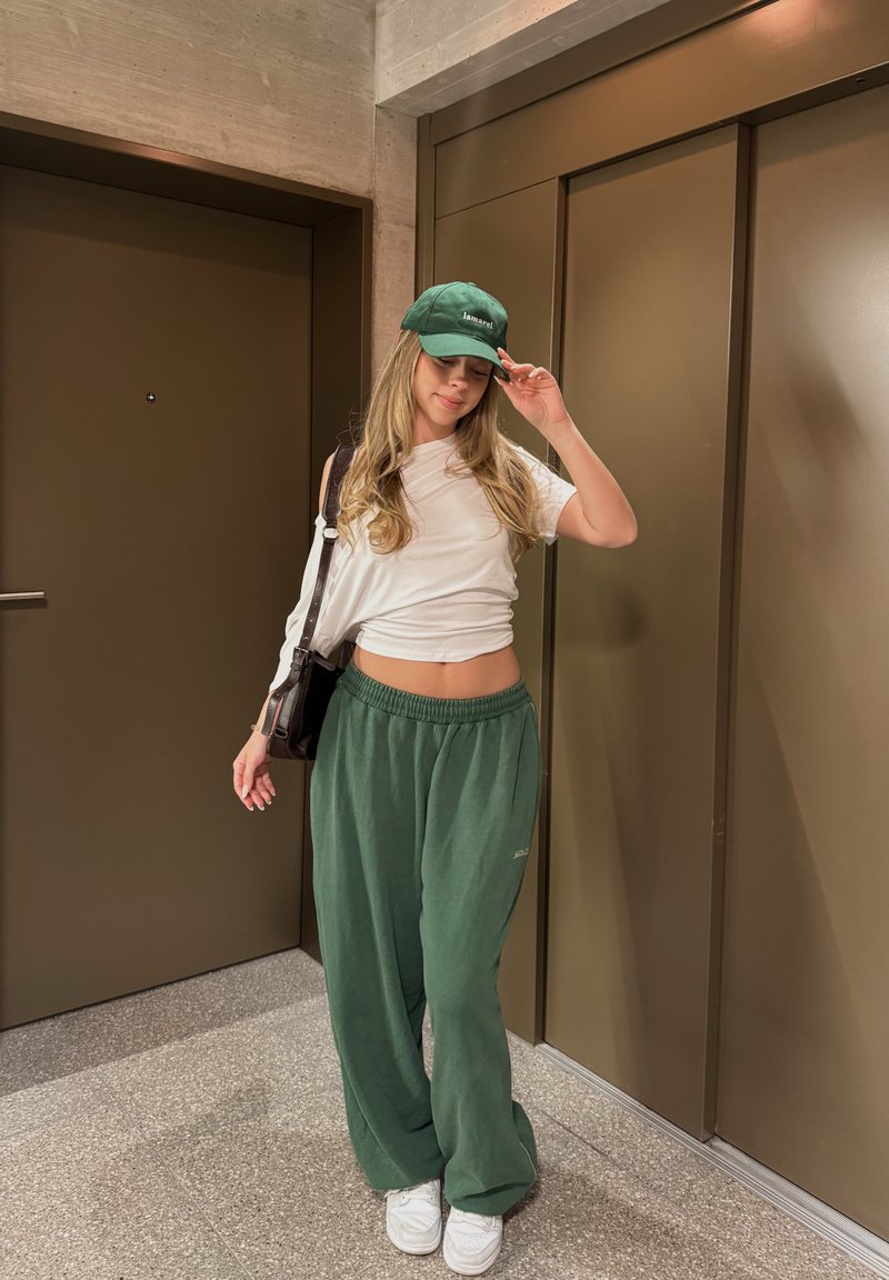 Junge Frau richtet grüne Kappe, trägt weißes bauchfreies Top, grüne Hose, weiße Sneakers, steht vor braunen Türen in Innenräumen.
