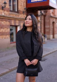 Veste en laine noire avec un col texturé, associée à une robe noire assortie. Tenant un sac à main compact noir avec des accessoires en argent.