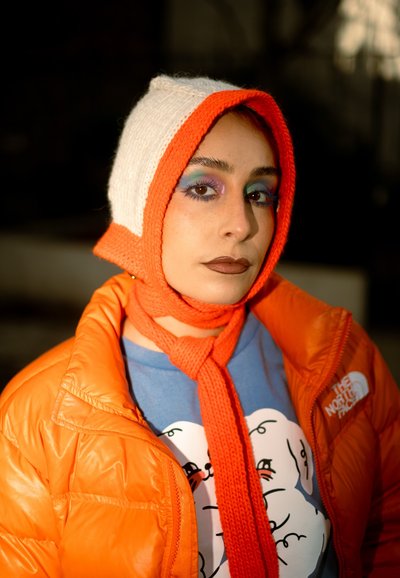 Persona con chaqueta acolchada naranja y gorro de punto naranja y blanco, con llamativa sombra de ojos azul y morada y lápiz labial marrón.