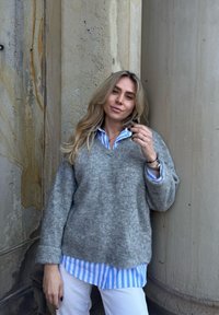 Pull gris en tricot sur une chemise à rayures bleues, associé à un pantalon blanc. Une femme s'appuie contre un mur en pierre texturée.