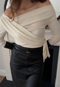 Γυναίκα που φοράει ένα κρεμ πουλόβερ τύπου off-shoulder, μαύρη δερμάτινη φούστα με λεπτή ζώνη και μαύρα γάντια, στέκεται σε εσωτερικό χώρο.