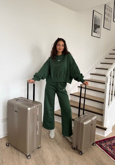 Mujer con chándal verde de pie en el interior sosteniendo dos maletas rígidas doradas, con una escalera y arte enmarcado al fondo.