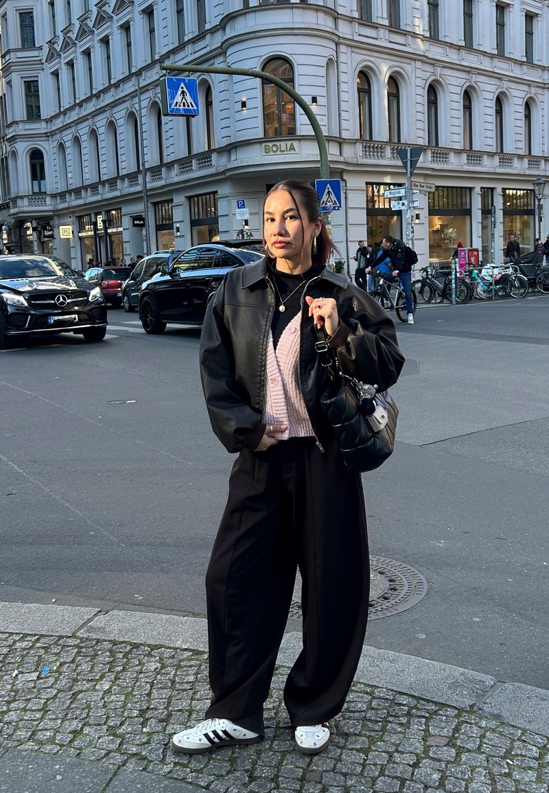 Svart läderjacka över en rosa stickad cardigan, vida svarta byxor och vita sneakers med svarta ränder. Håller en svart handväska.