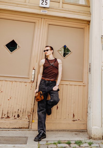 Top marrón sin mangas con un detalle de arnés, jeans negros de cintura alta, zapatos negros y un bolso cruzado marrón, todo ello frente a puertas pálidas.