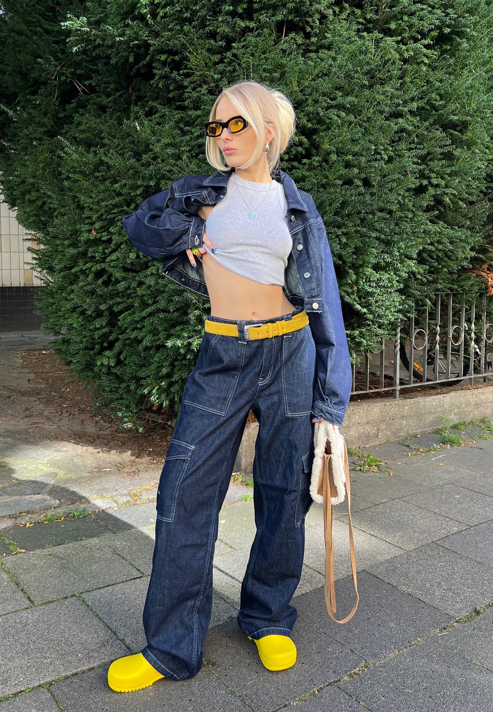 Denim jakna in široke hlače z visokim pasom v modri barvi z rumenim pasom; siva kratka majica; svetlo rumeni clogs; bež torba; ozadje z zelenjem.