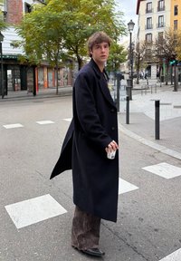 Dlhý tmavý kabát, hnedé kordurové nohavice a čierne topánky. Drží šálku, stojí na priechode pre chodcov s mestským pozadím a stromami.