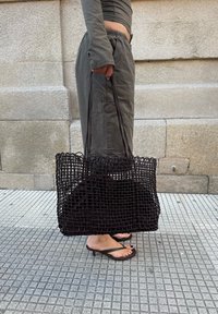 Bolso de mano negro tejido con asas largas, presenta un diseño de celosía abierta y un suave forro interior, posicionado sobre un fondo de piedra.