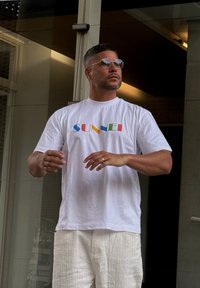 White T-shirt featuring a vibrant "SUNNEI" logo, paired with textured white shorts. Round sunglasses complete the casual ensemble.