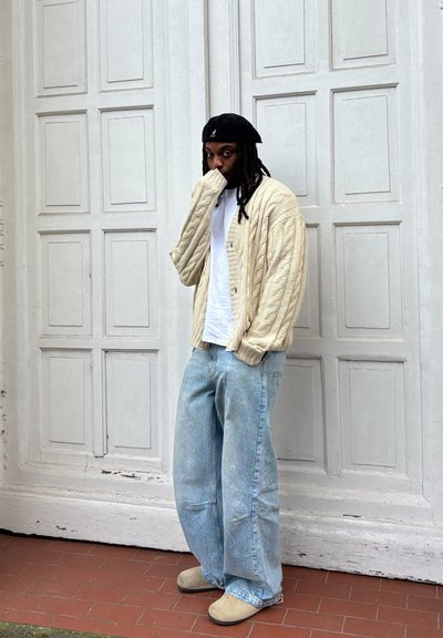 Cárdigan de punto con trenzas crema sobre una camisa blanca, combinado con unos vaqueros sueltos de color azul claro y unos zapatos beige sin cordones, todo ello frente a una pared blanca texturizada.