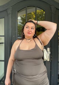 Καφέ αμάνικο φόρεμα με εφαρμοστή σιλουέτα και συγκεντρωμένη λεπτομέρεια, αξεσουαρισμένο με ένα ασημένιο κολιέ σε σχήμα καρδιάς και μια ανοιχτόχρωμη τσάντα.