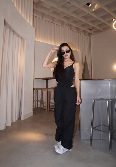 Bodysuit negro con tirantes finos y cortes laterales, combinado con pantalones cargo negros sueltos. Zapatillas blancas y unas pequeñas gafas de sol negras completan el look.