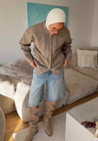 Veste en polaire marron avec fermeture à boutons, short en denim bleu clair et bottes hautes beige. Lunettes portées, foulard blanc sur la tête. Mobilier doux en arrière-plan.